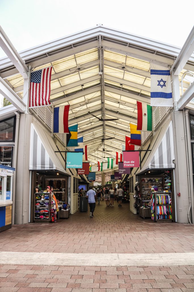 A foto mostra uma das ruas do shopping Bayside Market em Miami. Tem bandeiras dos países penduradas e as lojas.